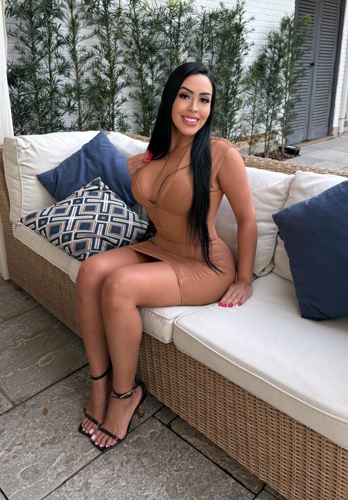 Massagista com longos cabelos pretos em um vestido bronzeado sentada em um sofá de vime ao ar livre com almofadas azuis e estampadas.